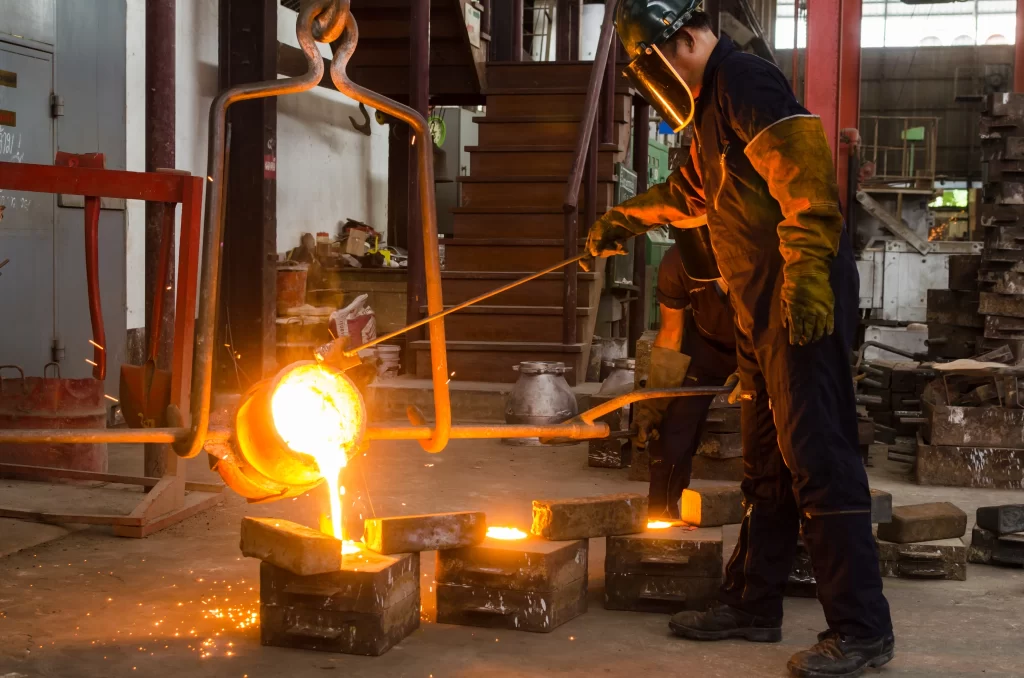 A worker in protective gear pours molten metal from a ladle into molds on a factory floor, creating glowing metal casts.