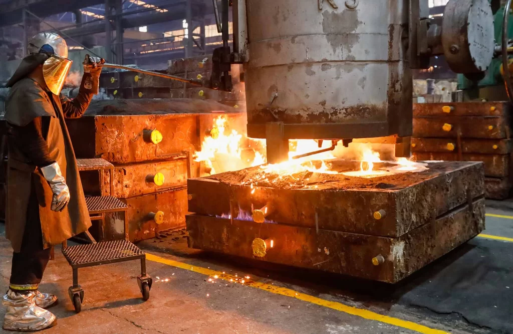 A worker in protective gear oversees molten metal being poured from a large machine into molds, sparks and flames visible.