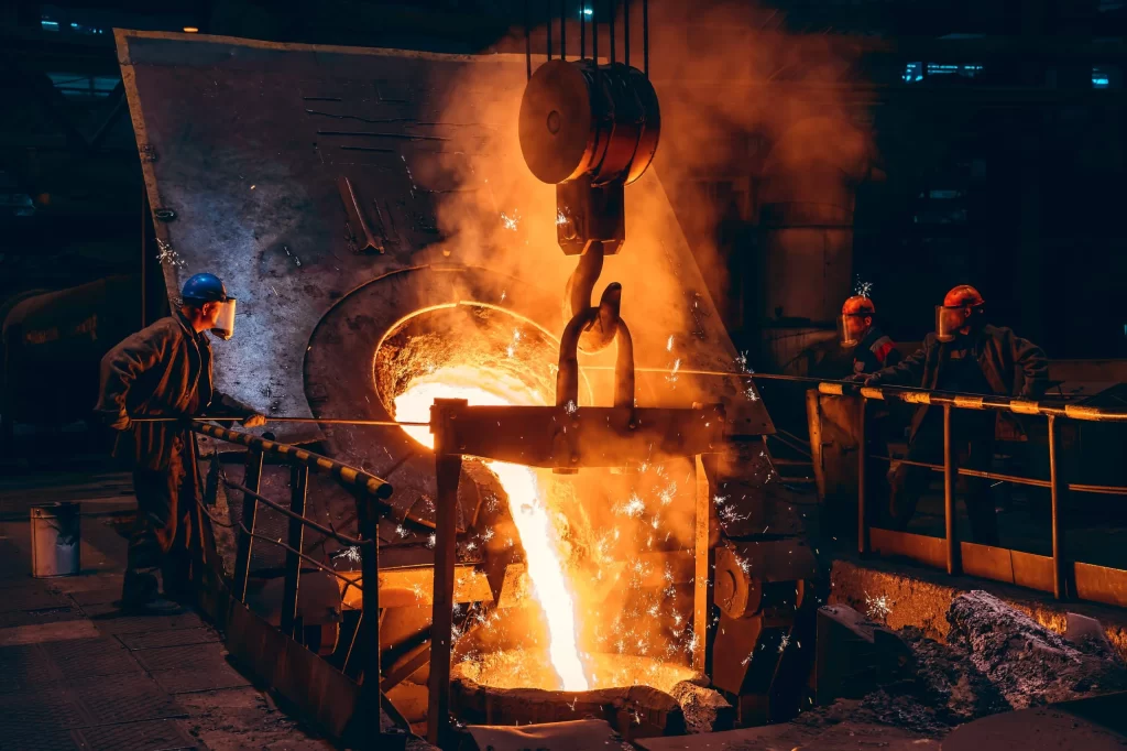 A metal foundry scene with workers in safety gear pouring molten metal from a large crucible, surrounded by smoke and intense heat.