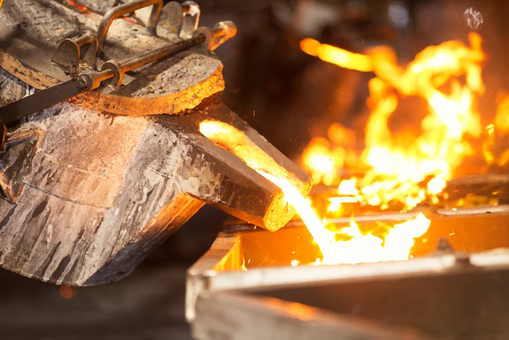 A molten metal is being poured from a ladle into a mold, surrounded by bright flames and industrial machinery in a foundry setting.