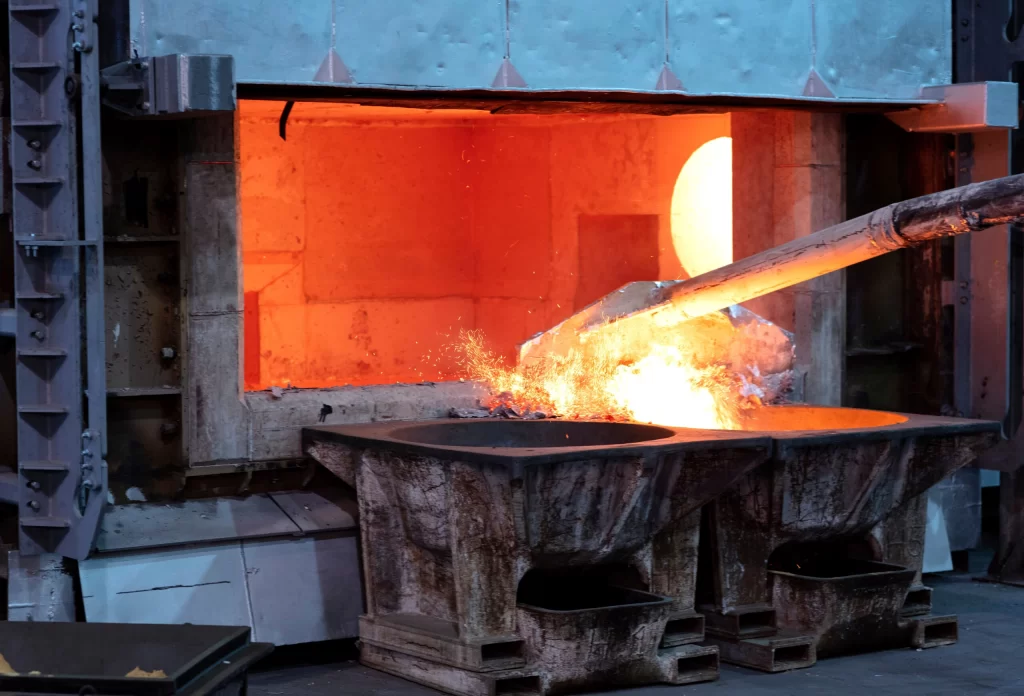 A molten metal pour from a furnace into two large containers, emitting bright sparks and intense orange glow in a foundry.