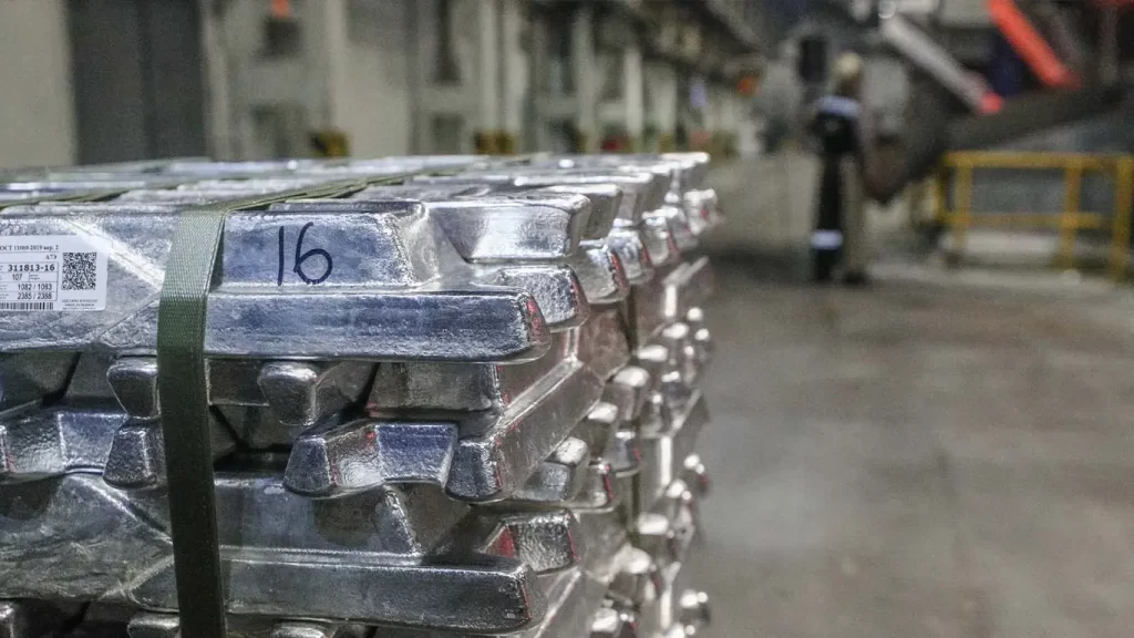 Stacks of aluminum ingots labeled with '16' in an industrial setting, with a worker in the background.