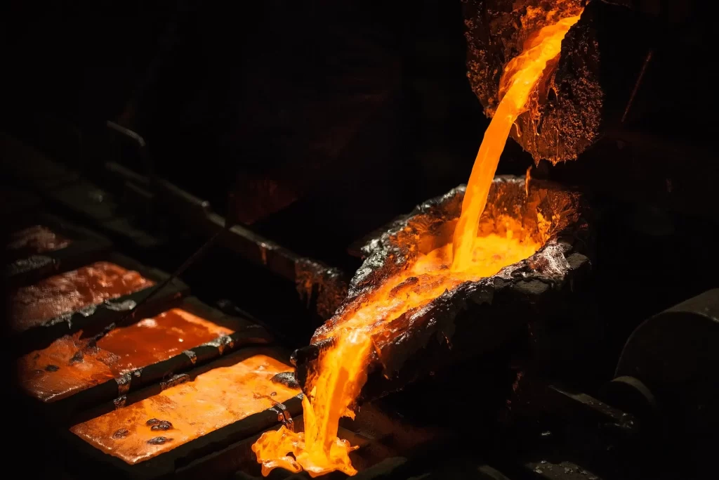 Molten metal pours from a furnace into molds, glowing bright orange against the dark background of a foundry.