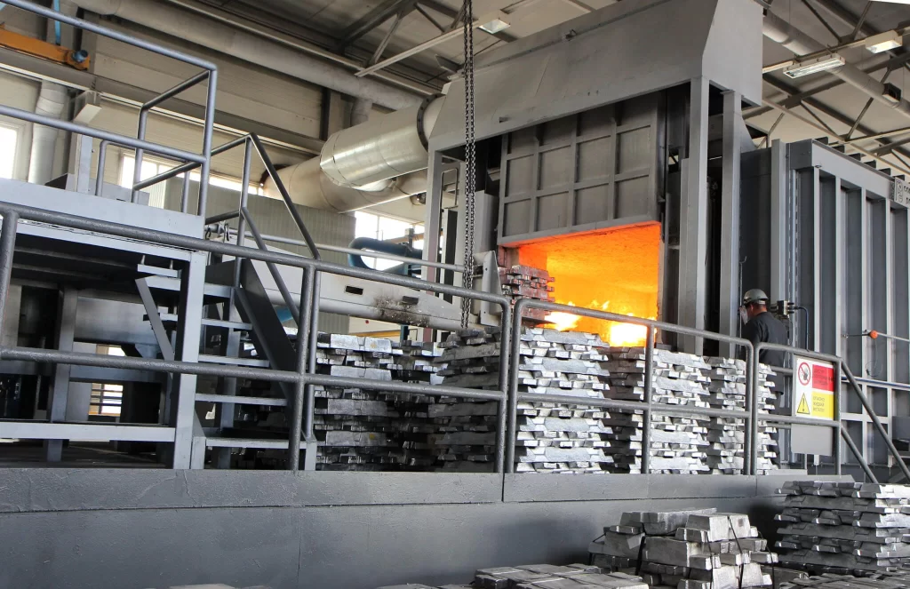 A metal foundry with an open furnace glowing orange; a worker stands nearby, surrounded by stacked metal bars on the platform.
