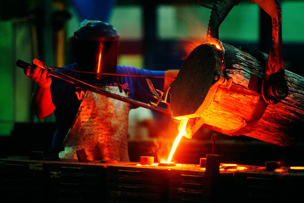 A worker in protective gear pours molten metal from a ladle into a mold, showcasing brass casting services in a dimly lit workshop with glowing orange light.