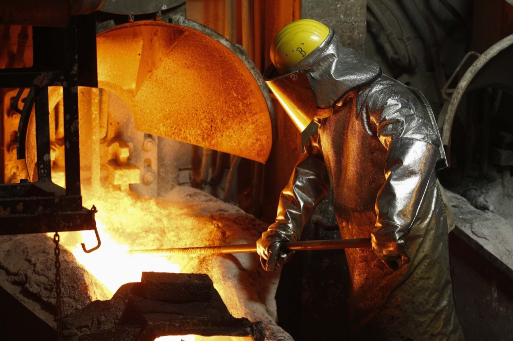 A worker in protective gear carefully maneuvers molten metal in a foundry during an aluminum bronze casting process, illuminated by the glowing furnace.