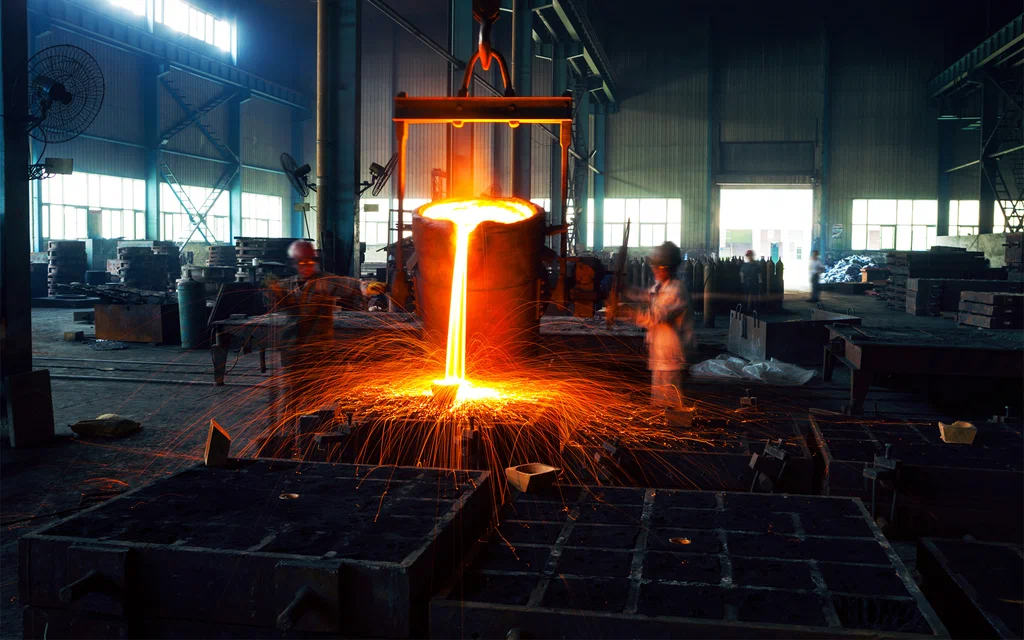 Workers in a foundry performing cast iron casting as they pour molten metal from a large ladle, producing bright sparks in an intensely lit industrial setting.