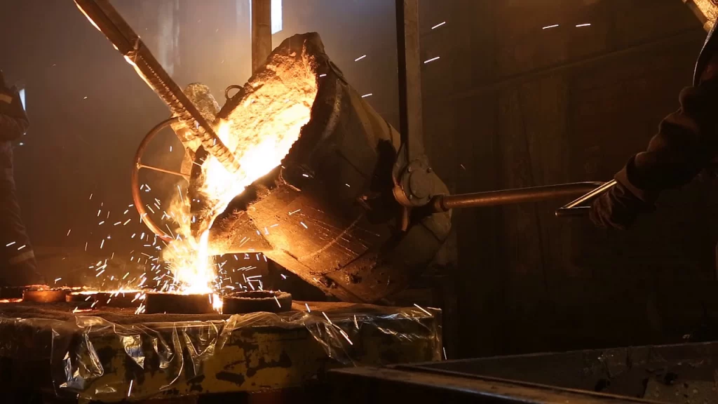 A worker pours molten metal from a large ladle, creating sparks, in a dimly lit foundry environment.
