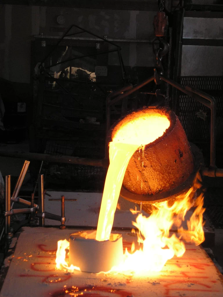 Molten metal being poured from a crucible into a mold, surrounded by flames, showcasing a dramatic metalworking process.