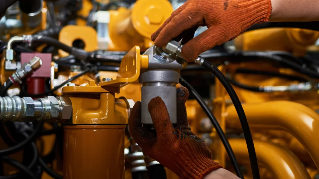 A worker in orange gloves performs hydraulic pressure testing by tightening a metal fitting on a yellow engine, surrounded by various pipes and machinery components.