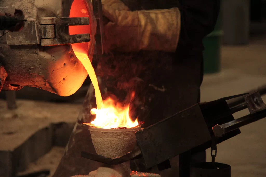 A gloved hand pours molten metal from a furnace into a stone mold, with vibrant flames and steam highlighting the intense heat process.