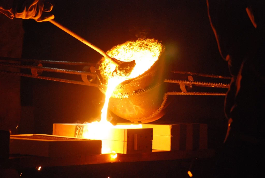 Molten metal is poured from a ladle during cast iron casting, creating bright sparks and glowing liquid in a dimly lit foundry environment.
