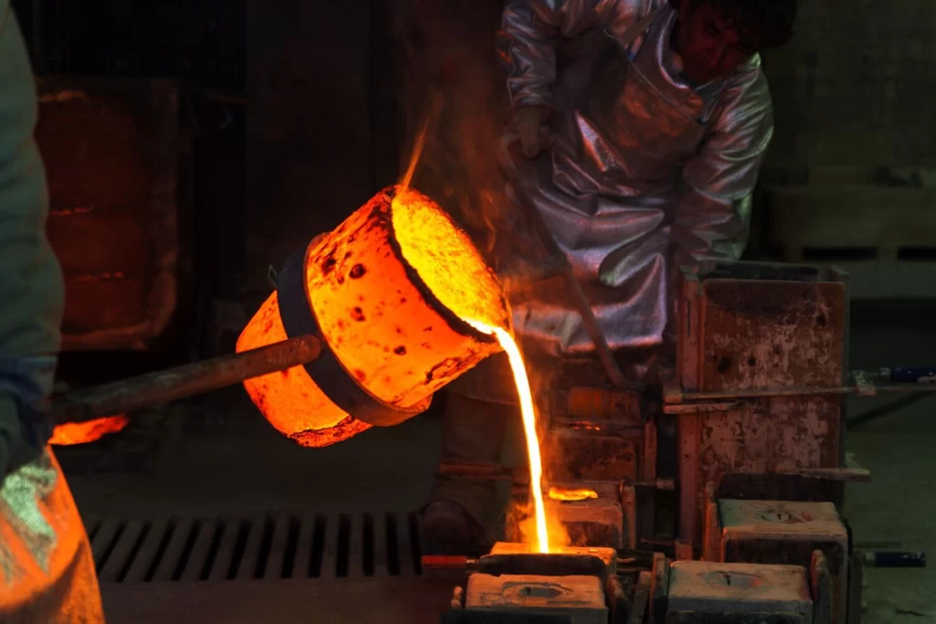 Molten metal pours from a ladle into a mold during cast iron casting, surrounded by steam and industrial equipment in a foundry setting.