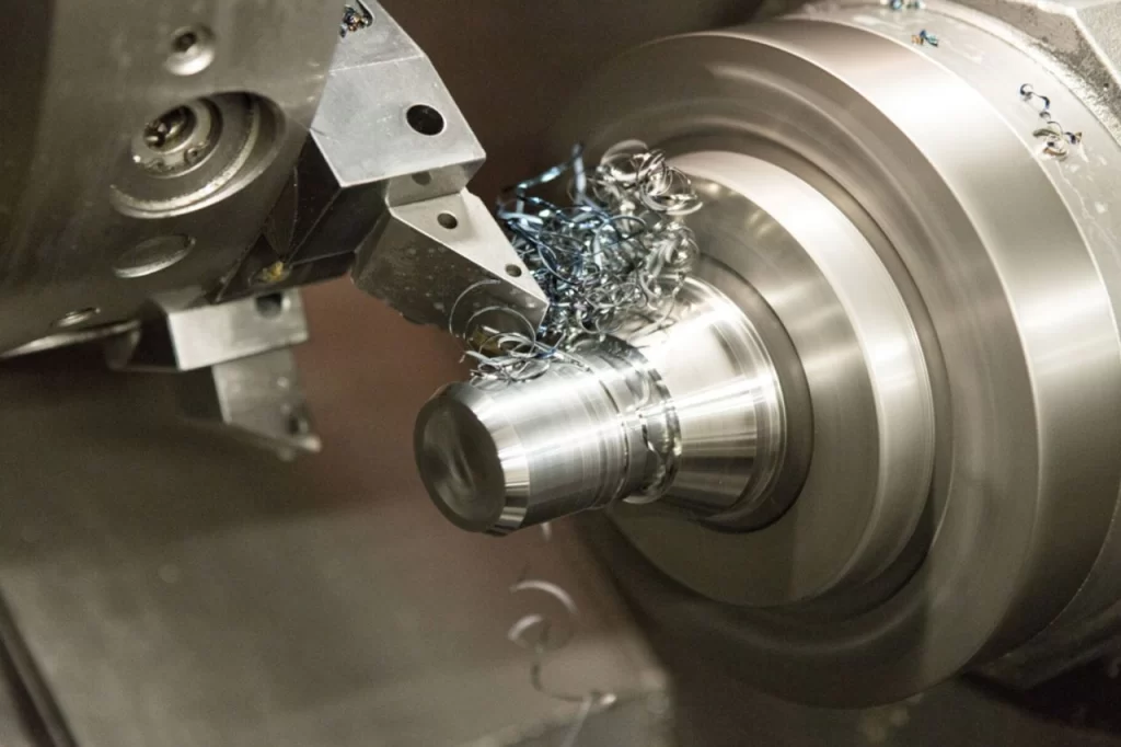 Close-up of lathe works on a metal lathe machining a cylindrical part, with metal shavings being cut away during the process.