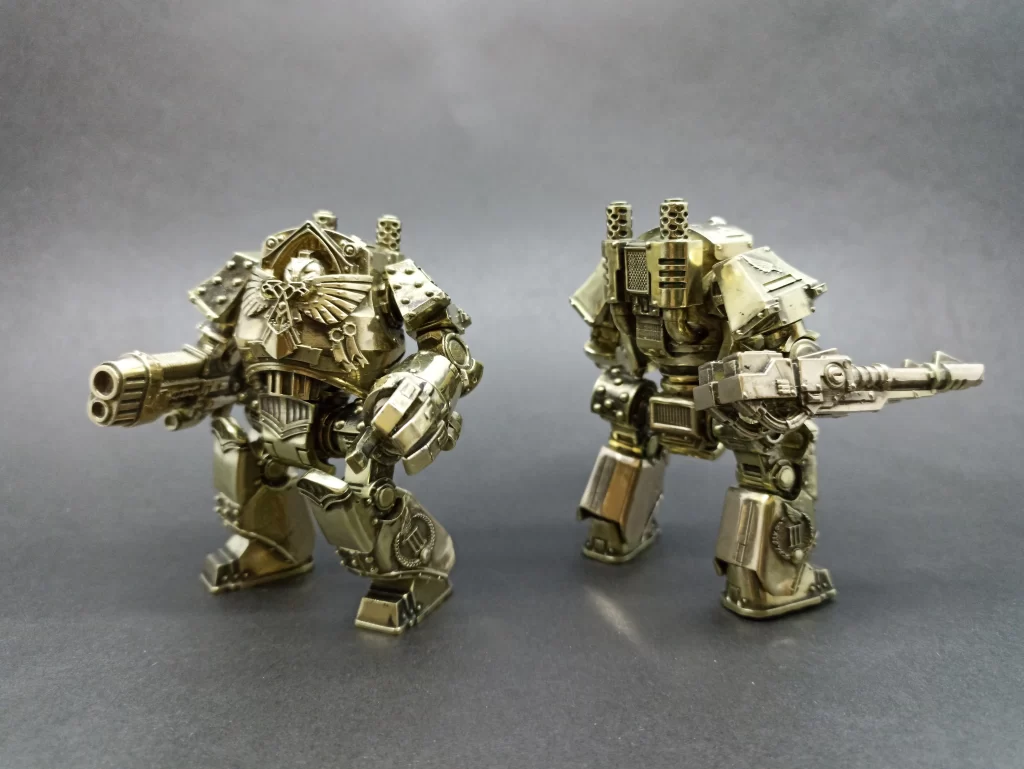Two metallic robot figures stand against a dark background, each equipped with heavy weapons and intricate detailing.