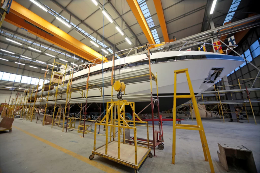 A large yacht is elevated on supports inside a spacious, well-lit industrial facility, surrounded by scaffolding and maintenance equipment.