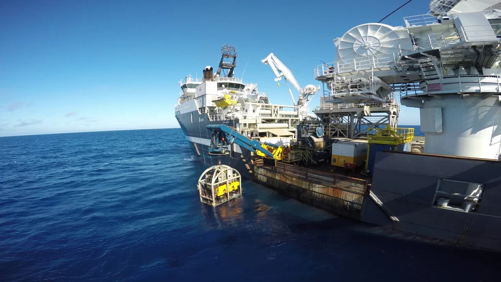 A deep-sea vessel equipped with machinery, positioned over blue waters, with a submersible vehicle being deployed.