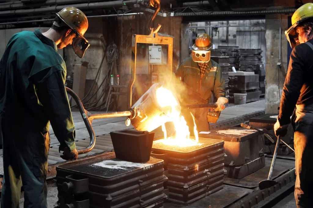 Three workers in safety gear pour molten metal into molds in a foundry, surrounded by machinery and glowing sparks. For Top UAE Metal Casting Services.