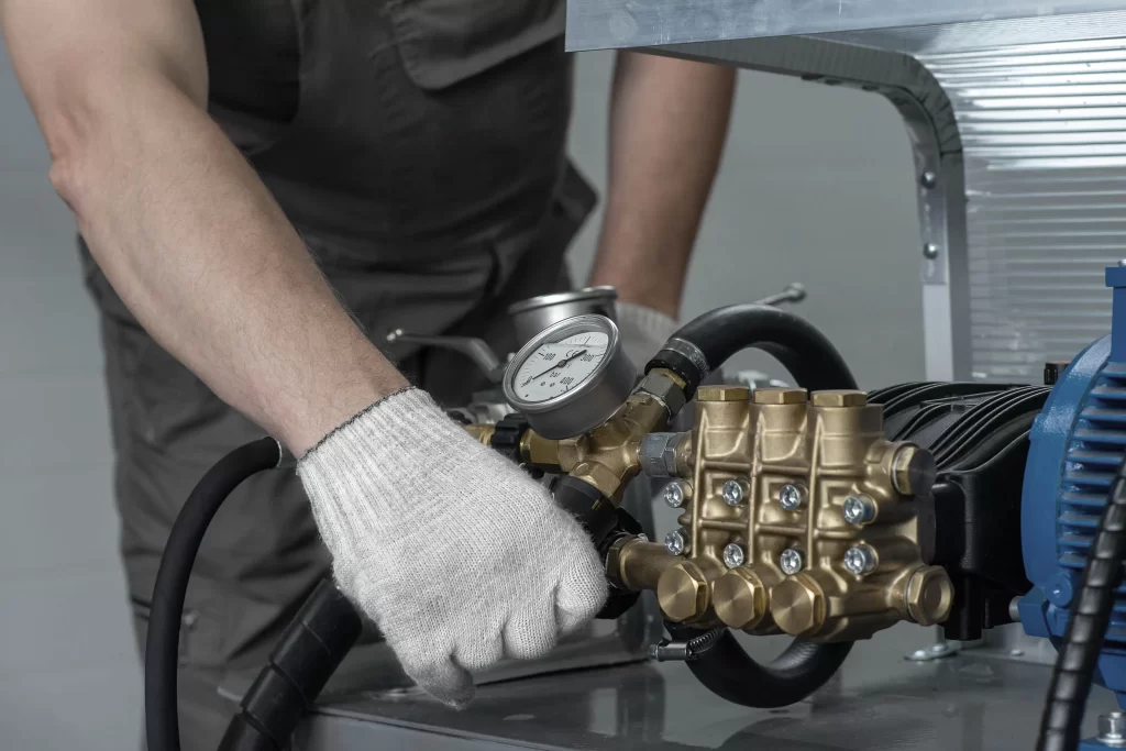 A technician in a gray jumpsuit, wearing a white glove, adjusts a pressure gauge on a brass pump during hydraulic pressure testing in a clean workshop setting.