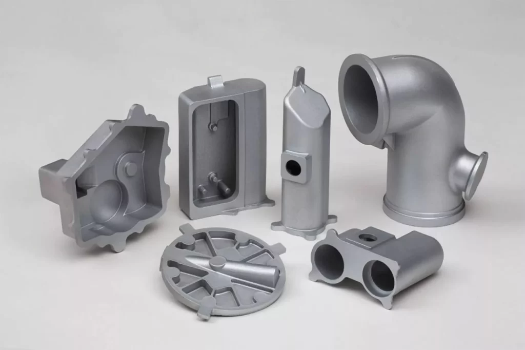 A collection of six metallic gray industrial metal parts, including a pipe elbow, casing, and various fittings, arranged aesthetically.