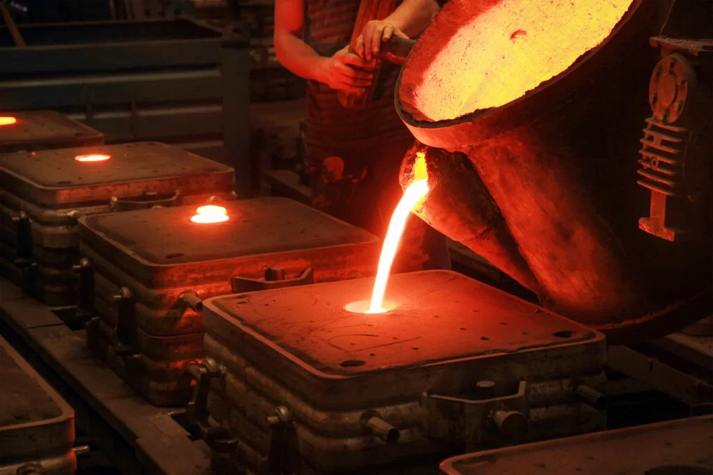 A worker pours molten metal from a ladle into molds in a foundry, surrounded by glowing metal and industrial equipment.