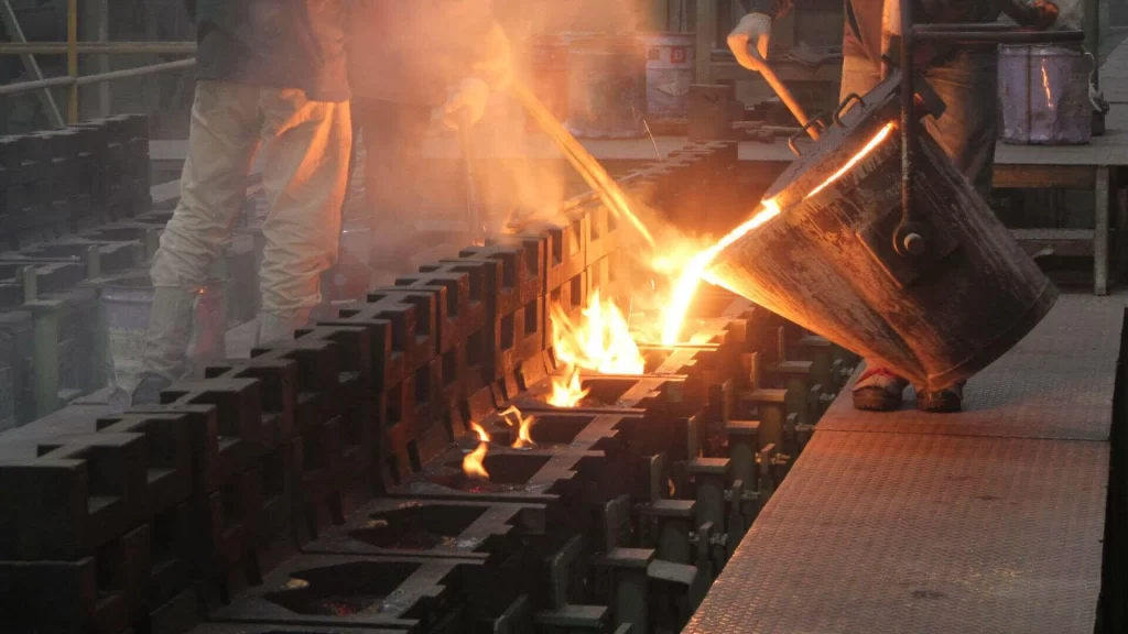 Workers pour molten metal into molds, surrounded by steam and flames in a busy foundry.