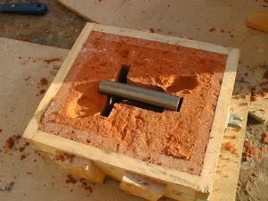 A wooden mold filled with orange clay, featuring a cylindrical metal pipe positioned in the center for casting purposes.