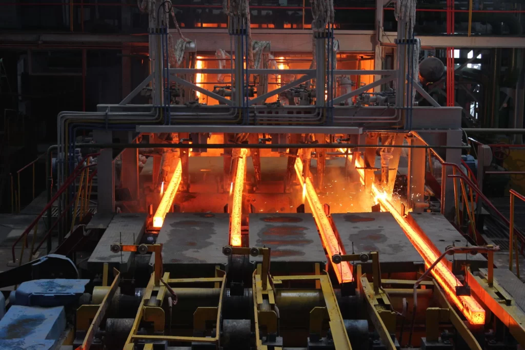 Industrial scene showing molten metal flowing from a furnace during an aluminum bronze casting process, illuminated by bright orange flames, with machinery and tracks in the foreground.