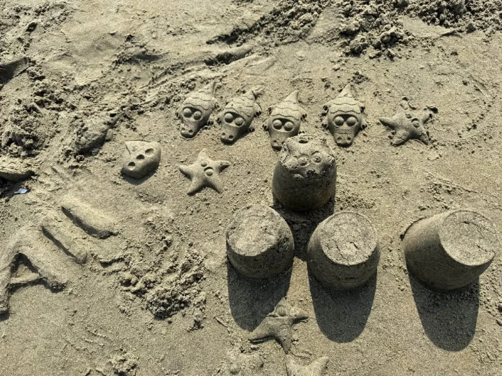 A sandy beach scene featuring playful sand sculptures including a crown, skulls, and starfish, with "ALL" written in the sand.