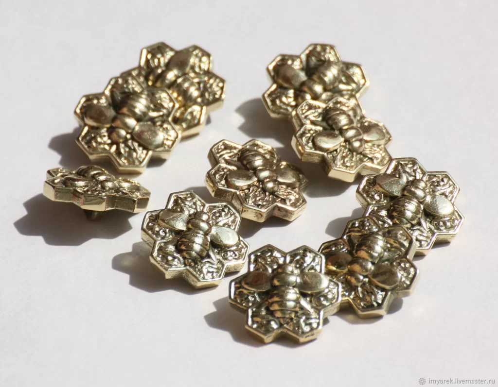 A collection of decorative metallic bee-shaped buttons arranged on a light background, showcasing intricate floral designs.