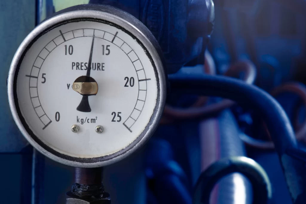 Close-up of a pressure gauge displaying readings in kg/cm², used for hydraulic pressure testing, set against blue industrial pipes.