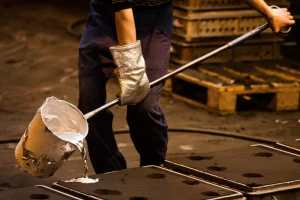 A worker in protective gloves pours molten metal from a ladle into molds in a manufacturing facility.