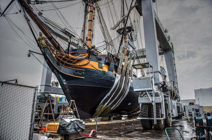 A large historic ship lifted by a travel lift in a boatyard, highlighting its rigging, sleek hull, and materials built for saltwater corrosion resistance.