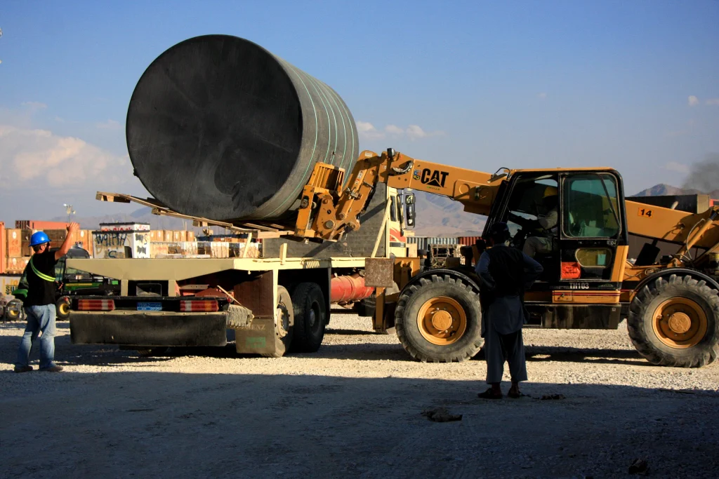 A construction site where a forklift loads a large metal pipe onto a flatbed truck under clear blue skies, symbolizing logistics in metal casting industries.