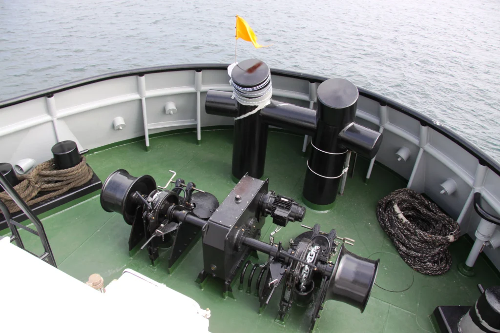 The image shows the bow of a ship with a green deck featuring marine casting equipment, mooring gear, and coiled ropes, with a yellow flag fluttering above.