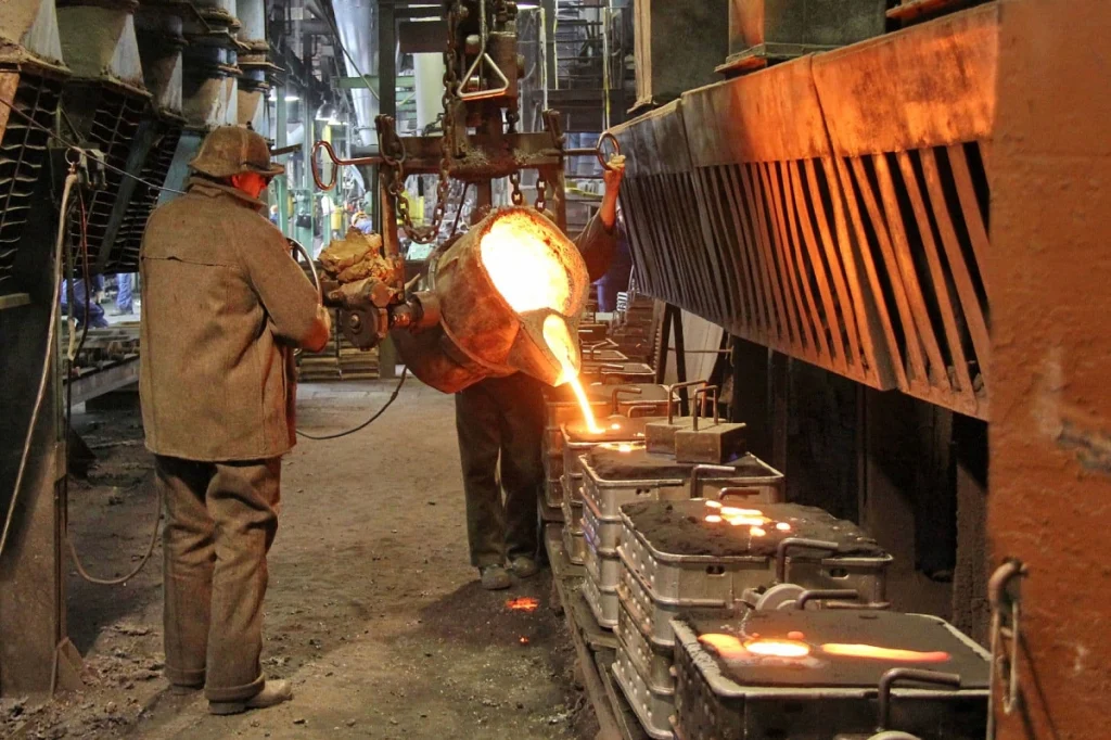 Two workers in a foundry pour molten metal into molds, illustrating the process of sand vs die casting in a glowing industrial setting.